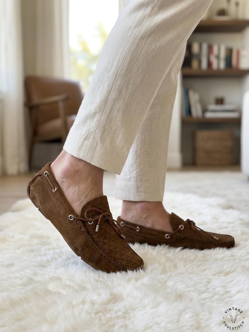 May include: Brown suede driving moccasins with a textured pattern and a leather tie detail. The shoes are worn with cream-colored pants on a white fluffy rug. The shoes have a classic, casual style.