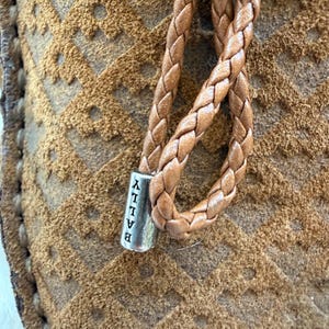 May include: Close-up of a brown suede shoe with a braided leather strap. The strap is attached to a silver metal piece with the brand name "BALLY" vertically printed on it. The suede has a geometric pattern.