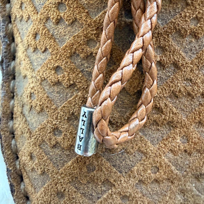 May include: Close-up of a brown suede shoe with a braided leather strap. The strap is attached to a silver metal piece with the brand name "BALLY" vertically printed on it. The suede has a geometric pattern.