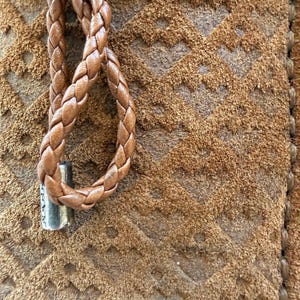 May include: Close-up of a brown leather item with a braided cord and a metal accent. The item has a textured surface with a geometric pattern. The braided cord is looped and attached to the item.
