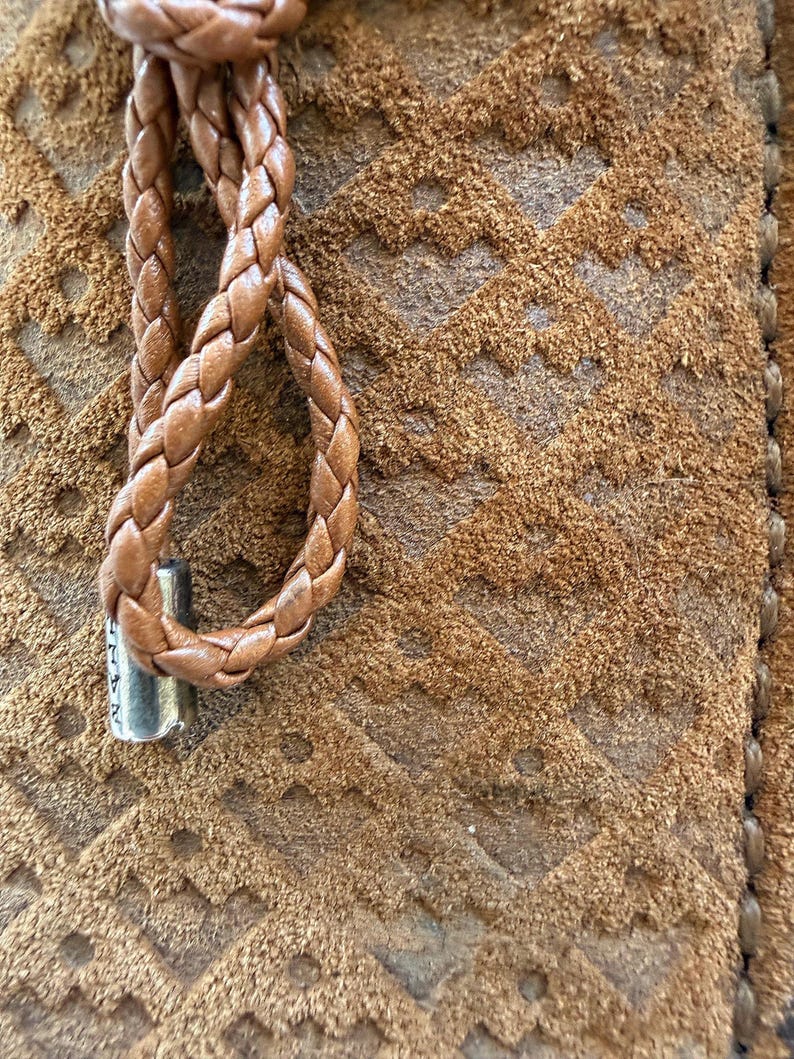 May include: Close-up of a brown leather item with a braided cord and a metal accent. The item has a textured surface with a geometric pattern. The braided cord is looped and attached to the item.