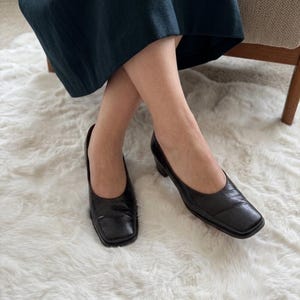 Può includere: Un paio di scarpe nere in pelle con punta quadrata e tacco basso. Le scarpe hanno una superficie strutturata. La persona indossa una gonna verde scuro. Le scarpe sono indossate da una persona seduta su un tappeto bianco e soffice.