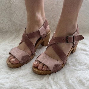 May include: A pair of dusty rose and mauve suede sandals featuring a wooden heel and sole. The footwear has crisscross straps across the top, a wide toe band, and an adjustable ankle strap with a metal buckle. Small metal studs secure the leather straps to the wooden base.