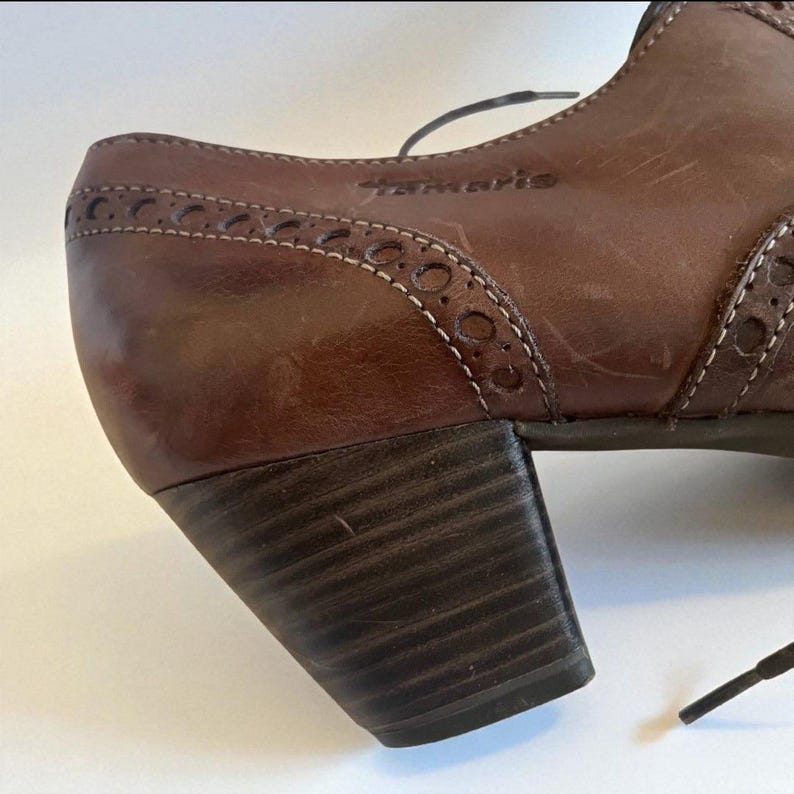 May include: Close-up of a brown leather shoe with a block heel. The shoe features decorative perforations along the edges and the brand name "tamaris" is visible. The heel is approximately 5 cm high.