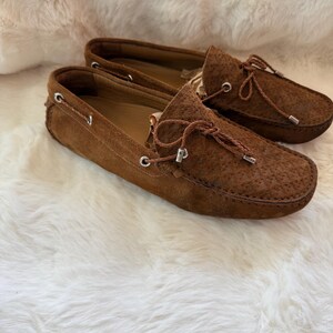 May include: A pair of brown suede loafers with decorative laces and metal eyelets. The shoes are displayed on a white, fluffy surface. The loafers have a textured design on the toe.