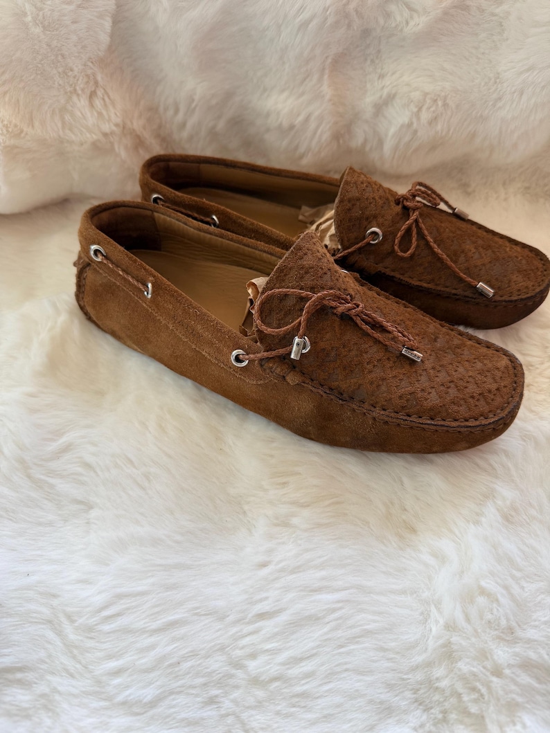 May include: A pair of brown suede loafers with decorative laces and metal eyelets. The shoes are displayed on a white, fluffy surface. The loafers have a textured design on the toe.