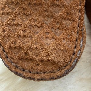 May include: Close-up of a brown suede shoe with a textured, diamond-patterned design. The shoe has a stitched border and a soft, worn appearance. The suede is a warm, earthy tone.