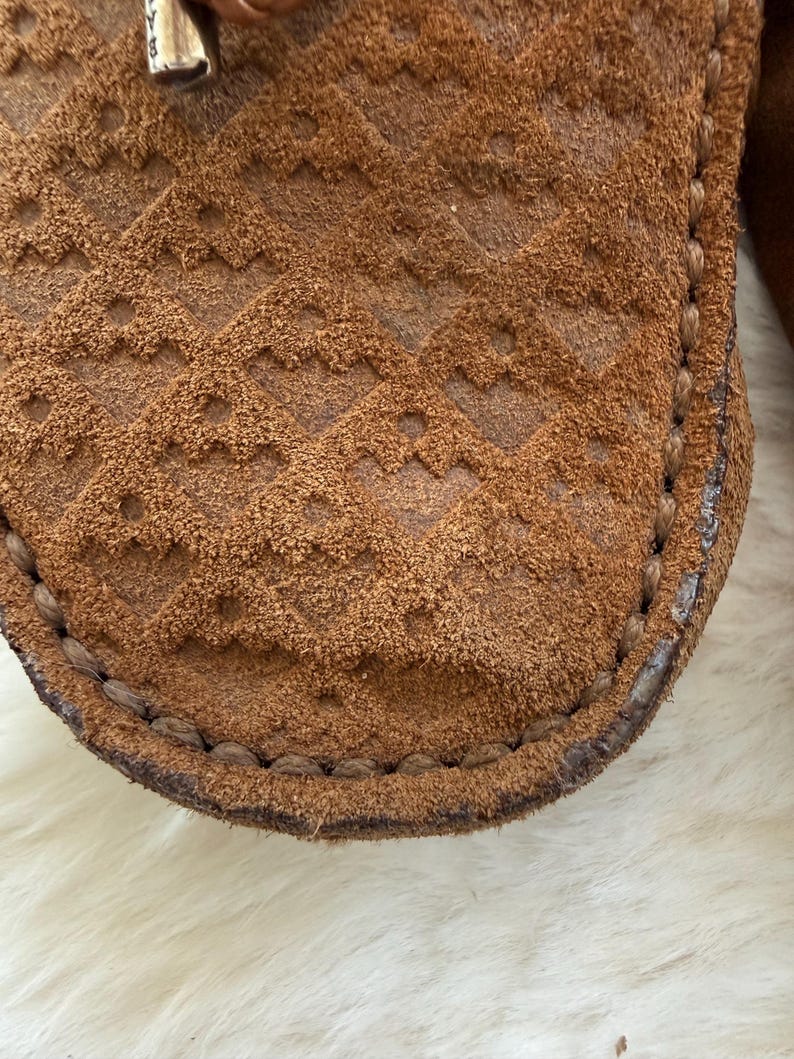 May include: Close-up of a brown suede shoe with a textured, diamond-patterned design. The shoe has a stitched border and a soft, worn appearance. The suede is a warm, earthy tone.