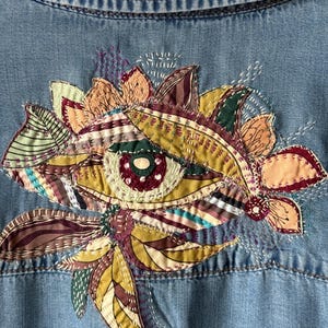 May include: A light blue denim shirt featuring an intricate, multi-colored embroidered eye design on the back. The embroidery incorporates various fabric patches in shades of olive green, burgundy, and cream, accented with detailed white stitching.
