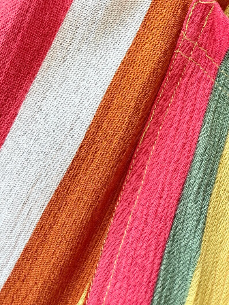 K&ouml;nnte beinhalten: Nahaufnahme eines farbenfrohen gestreiften Stoffes. Der Stoff weist kr&auml;ftige Streifen in Rot-, Orange-, Wei&szlig;-, Rosa-, Gr&uuml;n- und Gelbt&ouml;nen auf. Die Textur des Stoffes ist sichtbar, mit einem leicht geknitterten Aussehen. Die N&auml;hte sind mit gelben N&auml;hten sichtbar.