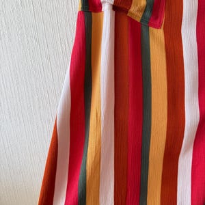 K&ouml;nnte beinhalten: Nahaufnahme eines Kleidungsst&uuml;cks mit vertikalen Streifen in Rot, Wei&szlig;, Orange, Gelb und Gr&uuml;n. Der Stoff hat ein strukturiertes, leicht gekr&auml;useltes Aussehen. Oben ist eine Tasche mit dem gleichen Streifenmuster zu sehen.
