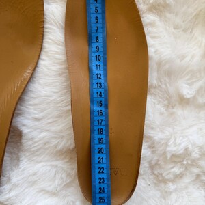 May include: A pair of brown leather insoles with a blue measuring tape alongside. The insole is approximately 11 inches long. The insoles are on a white, fluffy surface. The insoles are new.