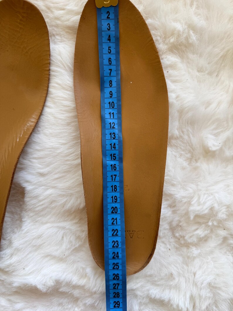 May include: A pair of brown leather insoles with a blue measuring tape alongside. The insole is approximately 11 inches long. The insoles are on a white, fluffy surface. The insoles are new.