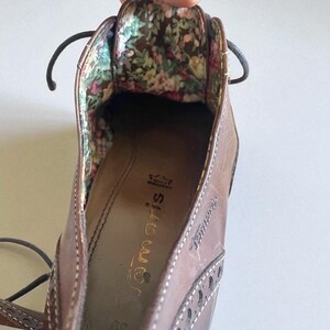 May include: Close-up of a brown leather shoe with a floral patterned interior. The shoe has a lace-up design and decorative stitching. The brand name "Tamaris" is visible on the insole.