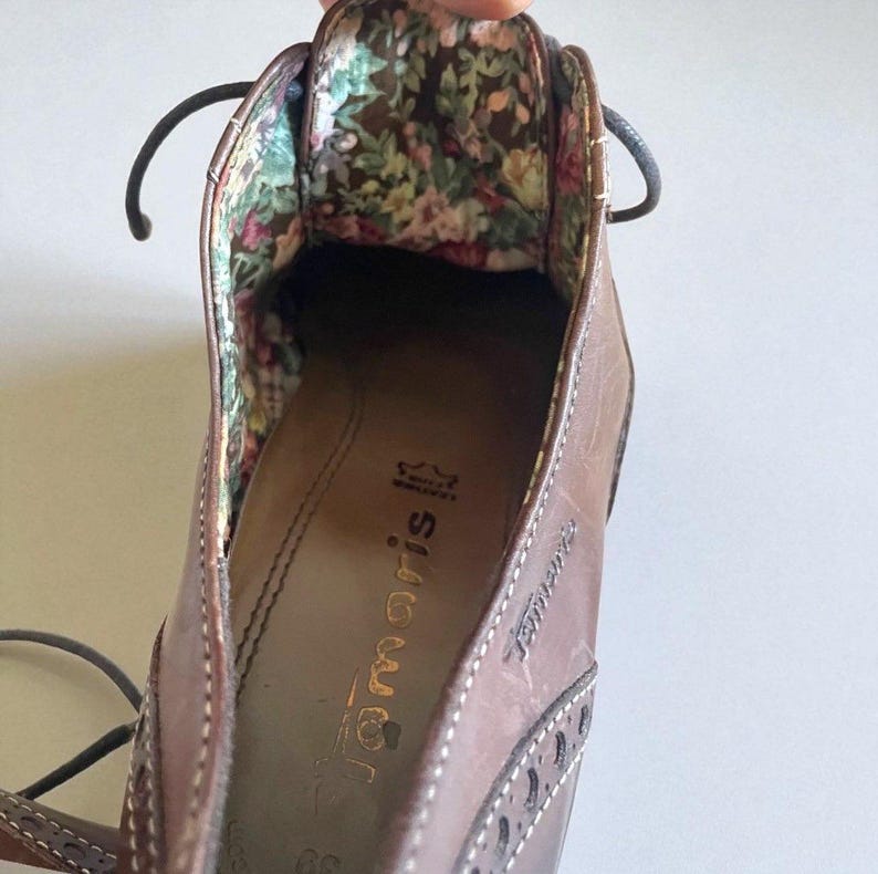 May include: Close-up of a brown leather shoe with a floral patterned interior. The shoe has a lace-up design and decorative stitching. The brand name "Tamaris" is visible on the insole.