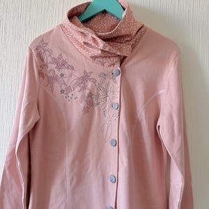 May include: A pale pink long-sleeved jacket with a high collar and button closure. The jacket has floral embroidery on the front and a contrasting dotted pattern on the collar. It is hanging on a teal hanger.