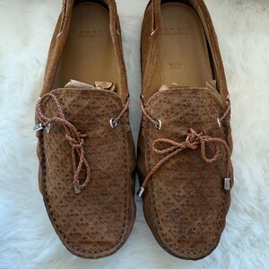 May include: Pair of brown suede loafers with intricate textured detailing. The shoes feature leather laces and silver-toned metal accents. The interior of the shoes is a lighter brown color, with the brand name visible. Made in Italy.