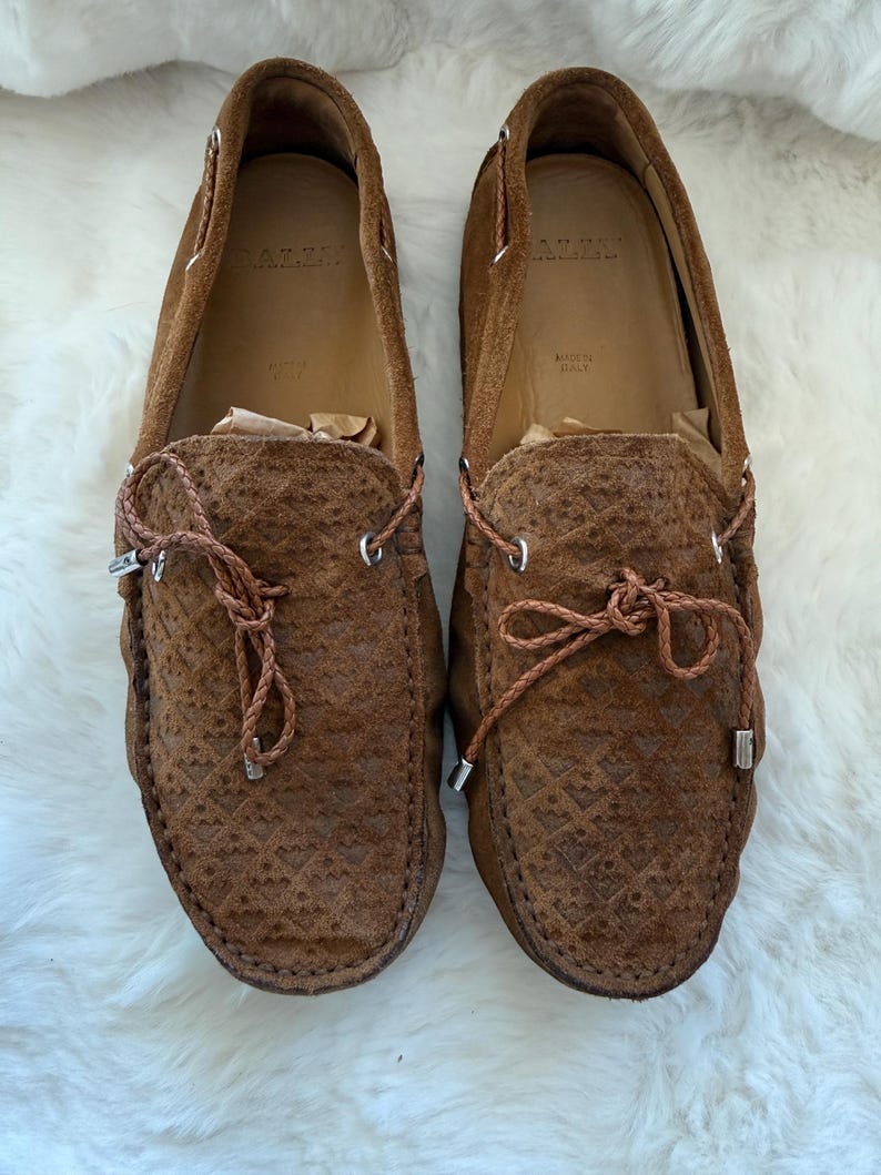 May include: Pair of brown suede loafers with intricate textured detailing. The shoes feature leather laces and silver-toned metal accents. The interior of the shoes is a lighter brown color, with the brand name visible. Made in Italy.