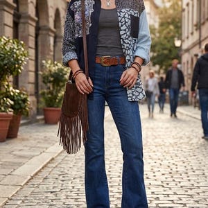 Puede incluir: Una camisa de botones tipo patchwork con paneles azul oscuro, azul claro y estampados sobre una camiseta sin mangas gris. El conjunto incluye vaqueros acampanados azul oscuro, un cinturón de cuero marrón con hebilla metálica, botas marrones y un bolso con flecos.