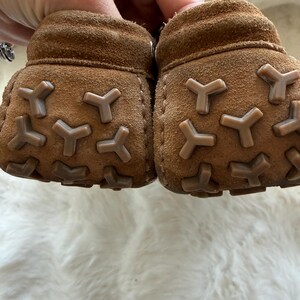 May include: A pair of brown suede baby booties with Y-shaped rubber soles. The soft leather shoes are designed for infants and feature a textured, non-slip sole for added grip. The booties are shown against a white, fluffy background.