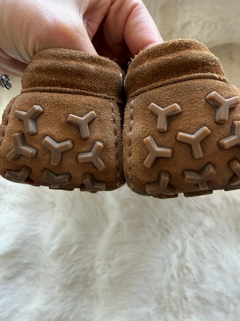 May include: A pair of brown suede baby booties with Y-shaped rubber soles. The soft leather shoes are designed for infants and feature a textured, non-slip sole for added grip. The booties are shown against a white, fluffy background.