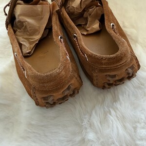 May include: A pair of brown suede loafers with a moccasin design. The shoes have a rounded toe, a rubber sole, and decorative stitching around the opening. The interior of each shoe is filled with brown paper. The shoes are on a white, fluffy surface.