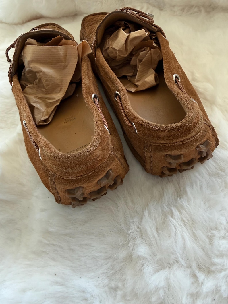 May include: A pair of brown suede loafers with a moccasin design. The shoes have a rounded toe, a rubber sole, and decorative stitching around the opening. The interior of each shoe is filled with brown paper. The shoes are on a white, fluffy surface.