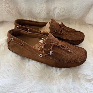 May include: A pair of brown suede loafers with a textured pattern. The shoes have a rounded toe, a leather lace detail, and a silver-toned metal accent. The shoes are displayed on a white, fluffy surface.