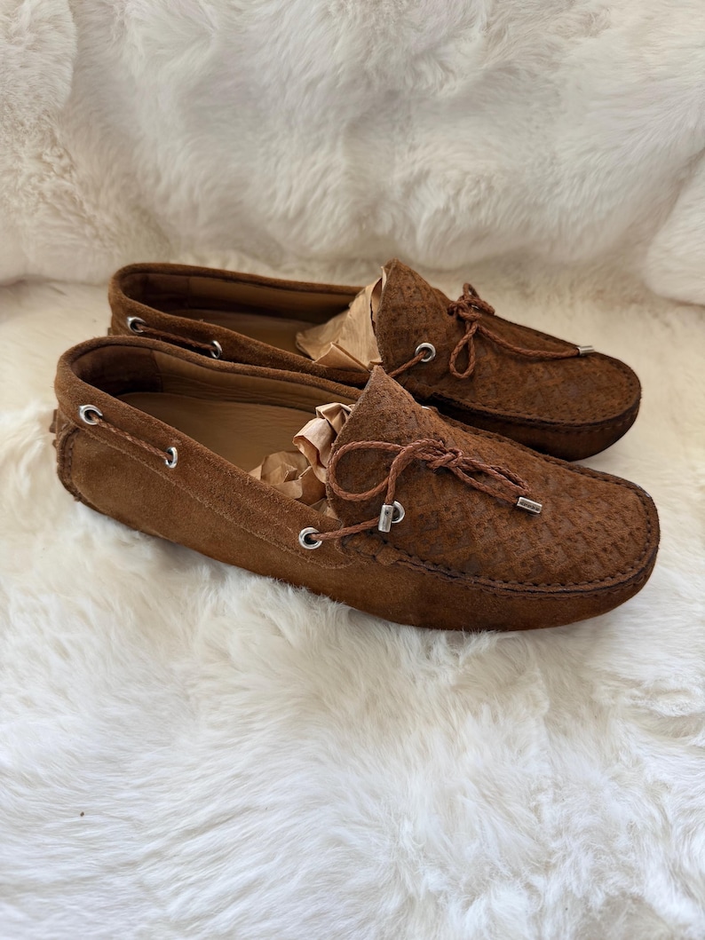 May include: A pair of brown suede loafers with a textured pattern. The shoes have a rounded toe, a leather lace detail, and a silver-toned metal accent. The shoes are displayed on a white, fluffy surface.