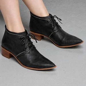 May include: Black leather ankle boots with a pointed toe and lace-up closure. The boots have a low, wooden heel and white stitching details. The soles are brown. The boots are shown on a gray surface.