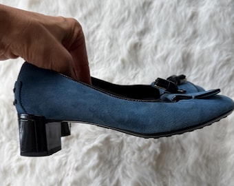 Puede incluir: Un zapato de tacón de ante azul con tacón de bloque negro y un lazo negro en la punta redondeada. El zapato presenta un borde negro a lo largo de la abertura y una suela negra texturizada. El diseño incluye un tacón bajo y acabado mate.