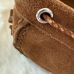 May include: Close-up of a brown suede loafer. The shoe features a braided leather detail threaded through a silver-toned metal eyelet. Visible stitching and a textured surface add to the shoe's design. The sole is a light brown color.