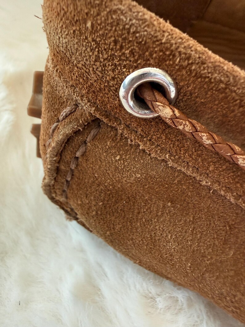 May include: Close-up of a brown suede loafer. The shoe features a braided leather detail threaded through a silver-toned metal eyelet. Visible stitching and a textured surface add to the shoe's design. The sole is a light brown color.