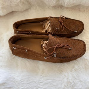 May include: A pair of brown suede loafers with a textured pattern. The shoes have a leather interior and a decorative bow on the top. The loafers are displayed on a white, fluffy surface.
