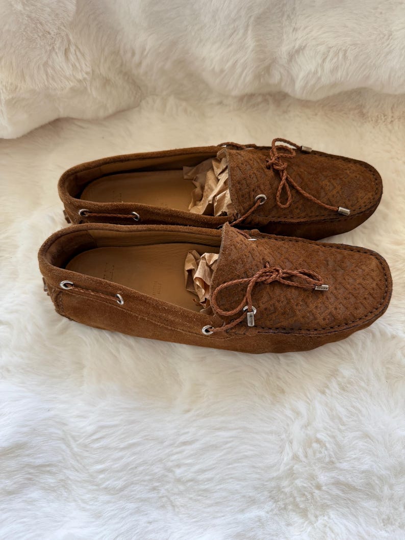 May include: A pair of brown suede loafers with a textured pattern. The shoes have a leather interior and a decorative bow on the top. The loafers are displayed on a white, fluffy surface.