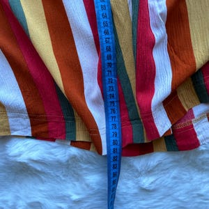 K&ouml;nnte beinhalten: Nahaufnahme eines farbenfrohen Stoffes mit Streifen und einem blauen Ma&szlig;band. Der Stoff hat vertikale Streifen in Rot, Orange, Gelb, Gr&uuml;n und Wei&szlig;. Das Ma&szlig;band zeigt Messungen von 147 bis 216 cm.