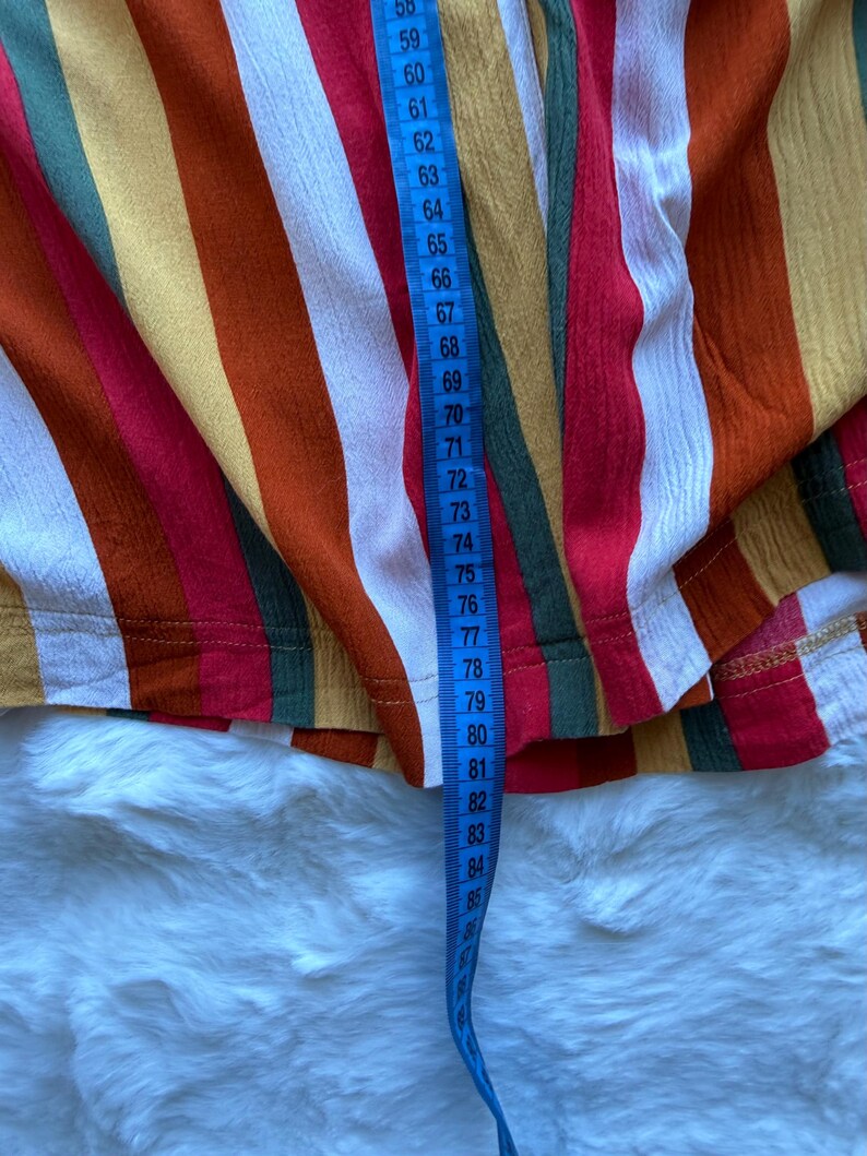 K&ouml;nnte beinhalten: Nahaufnahme eines farbenfrohen Stoffes mit Streifen und einem blauen Ma&szlig;band. Der Stoff hat vertikale Streifen in Rot, Orange, Gelb, Gr&uuml;n und Wei&szlig;. Das Ma&szlig;band zeigt Messungen von 147 bis 216 cm.