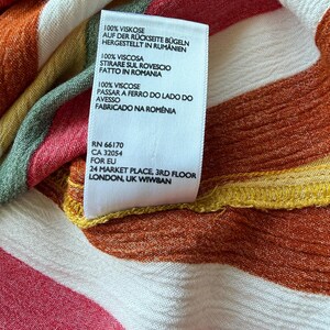 K&ouml;nnte beinhalten: Nahaufnahme eines Kleidungsst&uuml;cketiketts mit Text in mehreren Sprachen, darunter Deutsch, Italienisch und Portugiesisch. Das Etikett befindet sich auf einem Stoff mit farbenfrohen Streifen in Rot-, Orange-, Gelb- und Wei&szlig;t&ouml;nen. Der Stoff besteht aus 100 % Viskose.