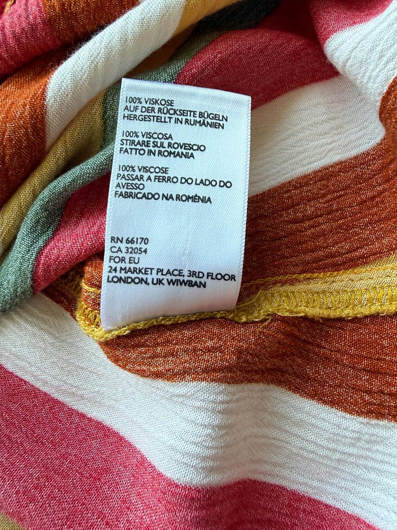 K&ouml;nnte beinhalten: Nahaufnahme eines Kleidungsst&uuml;cketiketts mit Text in mehreren Sprachen, darunter Deutsch, Italienisch und Portugiesisch. Das Etikett befindet sich auf einem Stoff mit farbenfrohen Streifen in Rot-, Orange-, Gelb- und Wei&szlig;t&ouml;nen. Der Stoff besteht aus 100 % Viskose.