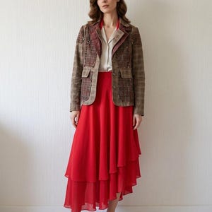 Könnte beinhalten: Ein langärmeliger Blazer mit braun-rotem Karomuster und Reverskragen, getragen über einem cremefarbenen Hemd und einem leuchtend roten, asymmetrischen Stufenrock aus Chiffon, kombiniert mit braunen Ledermokassins.