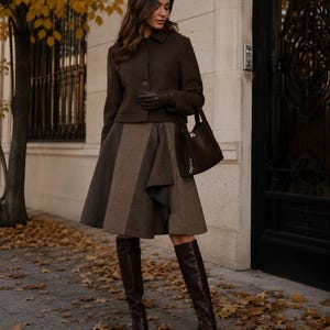 Puede incluir: Una mujer con un conjunto marrón, que incluye una chaqueta, falda, guantes y botas altas hasta la rodilla. La falda tiene un diseño de paneles único. El atuendo se combina con un bolso marrón. El fondo presenta un edificio con una puerta oscura y hojas de otoño.