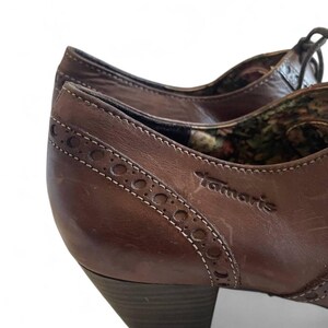 May include: Close-up of brown leather shoes with a low heel. The shoes feature decorative stitching and the brand name "Tamaris". The interior lining has a floral pattern.