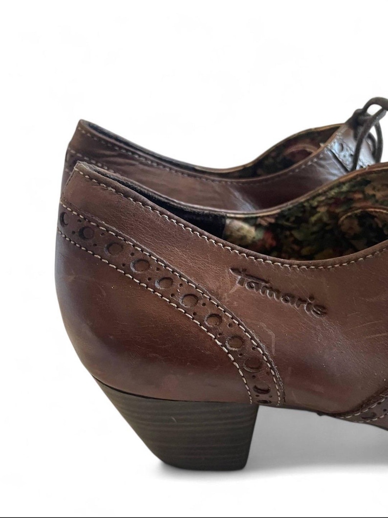 May include: Close-up of brown leather shoes with a low heel. The shoes feature decorative stitching and the brand name "Tamaris". The interior lining has a floral pattern.