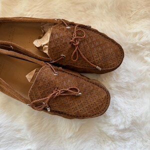 May include: A pair of brown suede loafers with a textured pattern. The shoes have a rounded toe and a leather tie detail. The shoes are displayed on a white, fluffy surface.