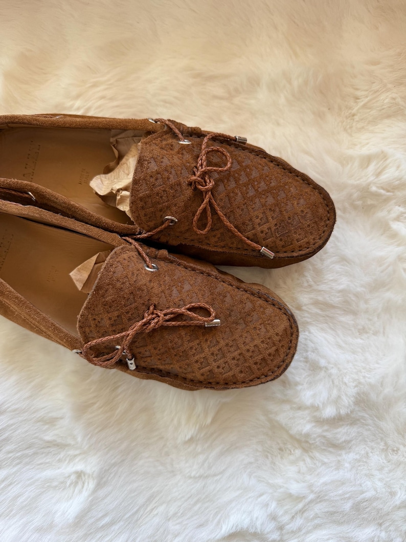 May include: A pair of brown suede loafers with a textured pattern. The shoes have a rounded toe and a leather tie detail. The shoes are displayed on a white, fluffy surface.