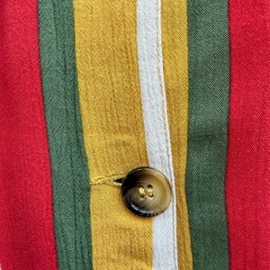 K&ouml;nnte beinhalten: Nahaufnahme eines Stoffes mit vertikalen Streifen in Rot, Gr&uuml;n, Gelb und Wei&szlig;. Ein brauner Knopf ist sichtbar, der am Stoff befestigt ist. Der Stoff scheint Teil eines Kleidungsst&uuml;cks zu sein.