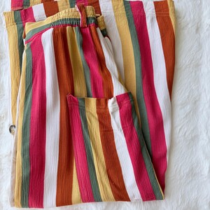 K&ouml;nnte beinhalten: Gefaltete Hose mit bunten Streifen. Die vertikalen Streifen umfassen Rot, Wei&szlig;, Gelb, Gr&uuml;n und Orange. Die Hose hat einen elastischen Bund und einen Knopfverschluss. Der Stoff hat ein strukturiertes Aussehen.