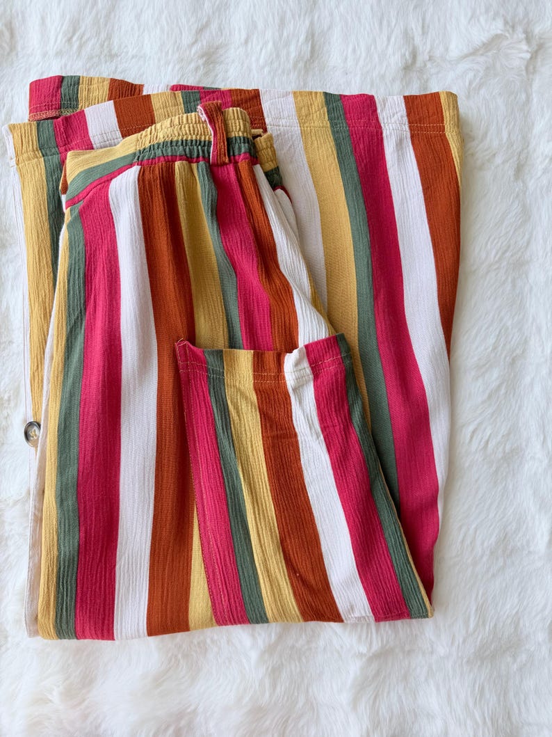 K&ouml;nnte beinhalten: Gefaltete Hose mit bunten Streifen. Die vertikalen Streifen umfassen Rot, Wei&szlig;, Gelb, Gr&uuml;n und Orange. Die Hose hat einen elastischen Bund und einen Knopfverschluss. Der Stoff hat ein strukturiertes Aussehen.