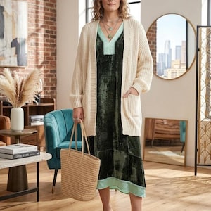 May include: A cream-colored open-front knit cardigan with two front pockets worn over a dark green velvet midi dress featuring a light green V-neck trim and hem. The outfit includes a woven straw tote bag and light green pointed-toe heels.