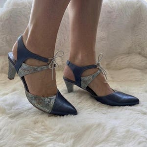 Op de afbeelding: Een paar marineblauwe en metallic zilveren schoenen met patroon, een puntige neus, een gesloten hiel en een vetersluiting bij de enkel. De schoenen hebben een blokhak van gemiddelde hoogte en een combinatie van effen marineblauw leer en gestructureerd zilverkleurig materiaal.